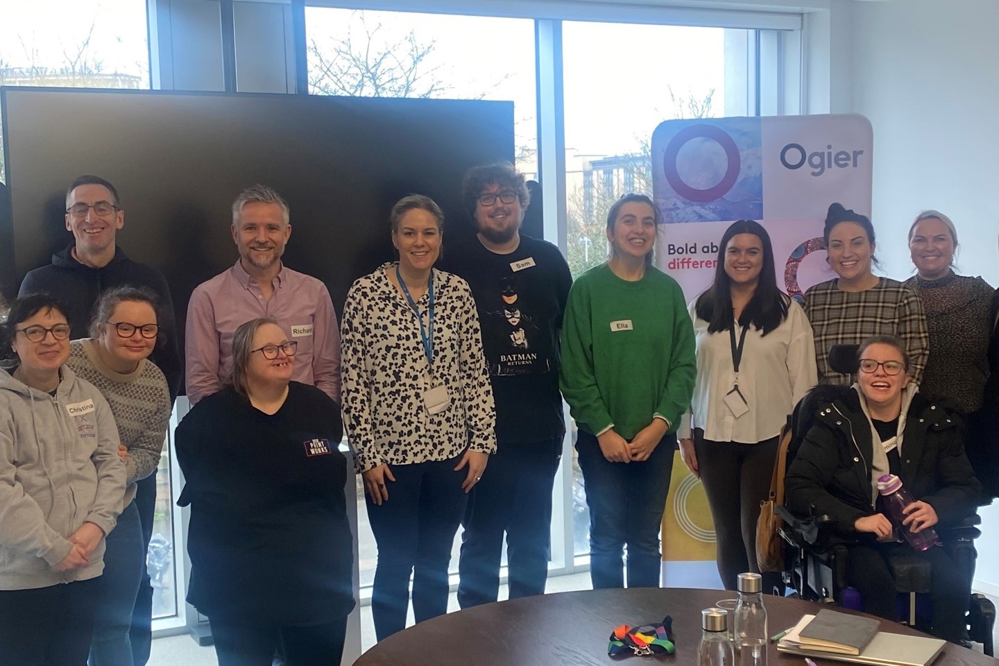 Ogier employees and six Beresford Street Kitchen crew members stand together in the Jersey office with BSK and Ogier banners behind them, celebrating partnership and workplace training initiative
