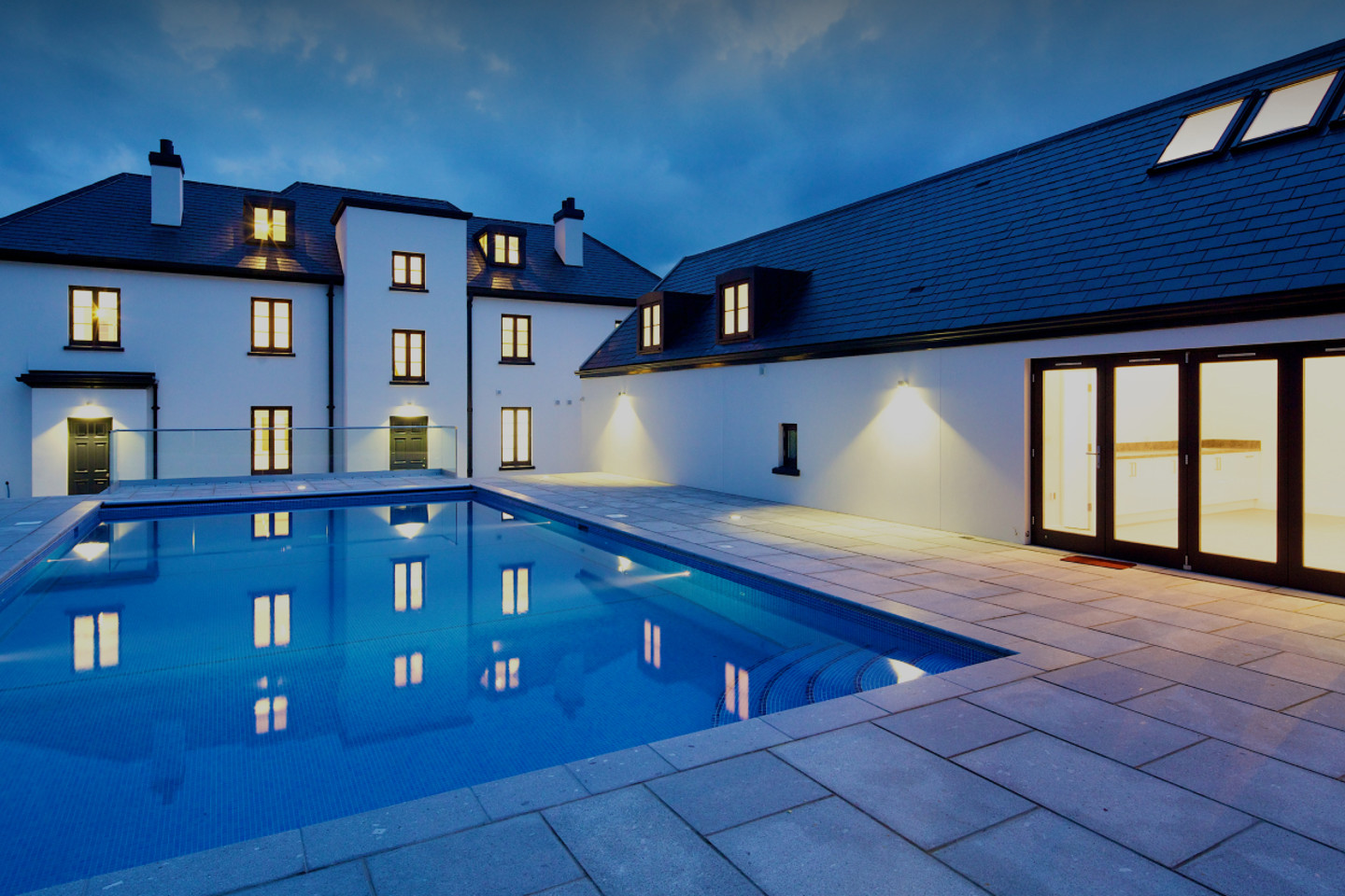 Large modern home with lit windows, slate roof and outdoor swimming pool at dusk